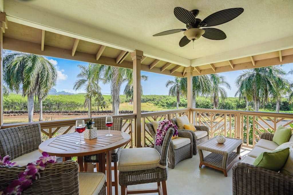 Een overdekt terras met rieten meubels, een plafondventilator en uitzicht op palmbomen en begroeiing.