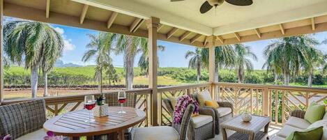 Een overdekt terras met rieten meubels, een plafondventilator en uitzicht op palmbomen en begroeiing.