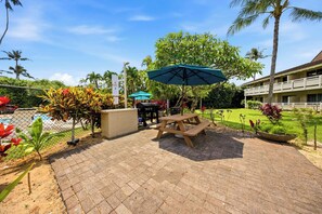 Ein Patio mit Picknicktisch und Grill, umgeben von tropischen Pflanzen und einem Pool.
