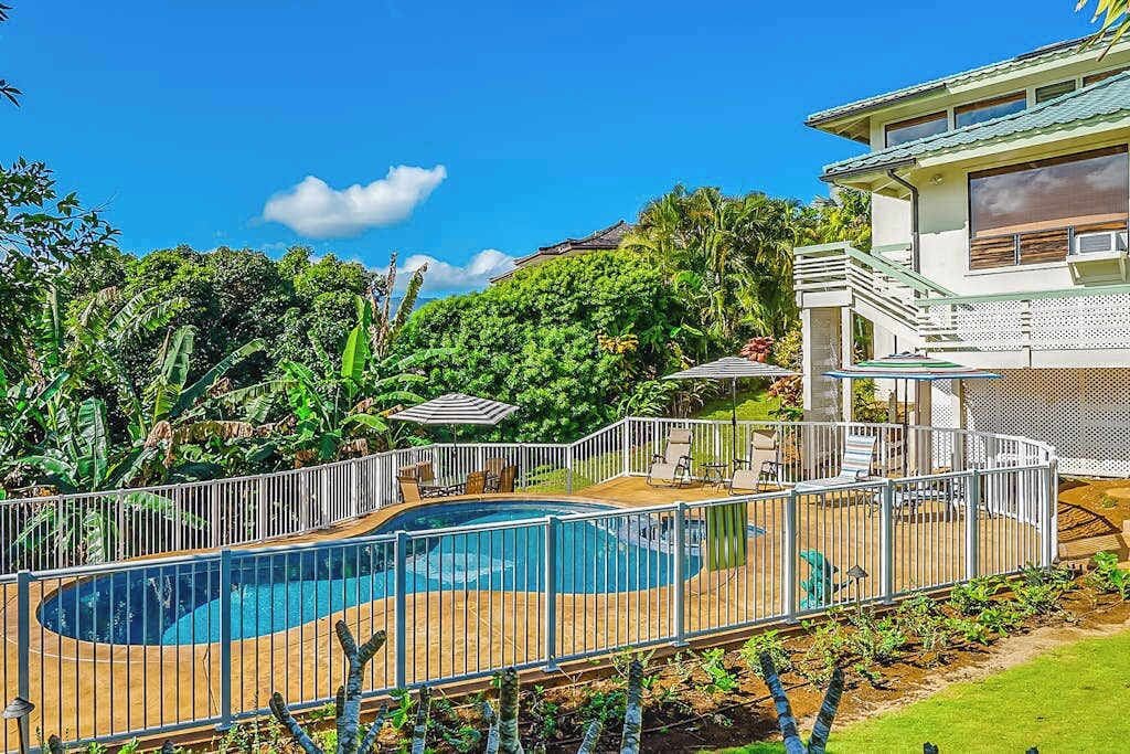Une piscine avec une terrasse en bois, entourée d’une clôture blanche, et une maison avec un toit vert en arrière-plan.