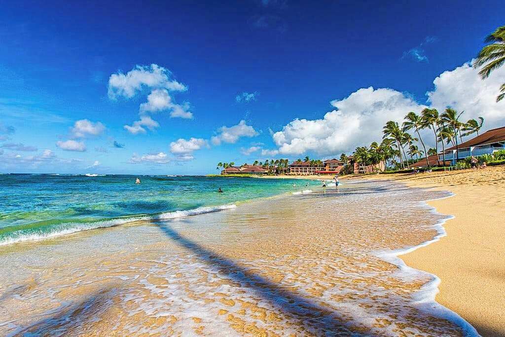 A beach with clear water, a sandy shore, and a resort with buildings and palm trees.