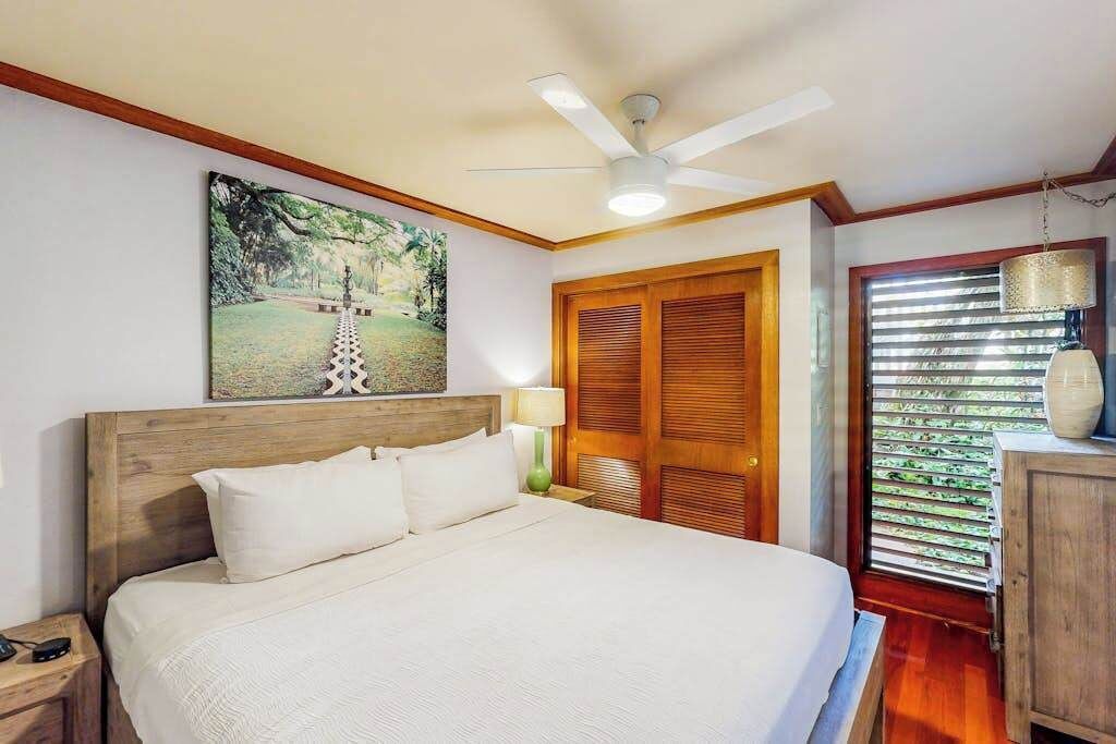 A bedroom with a bed, a ceiling fan, a painting, a wooden door, and a window with blinds.