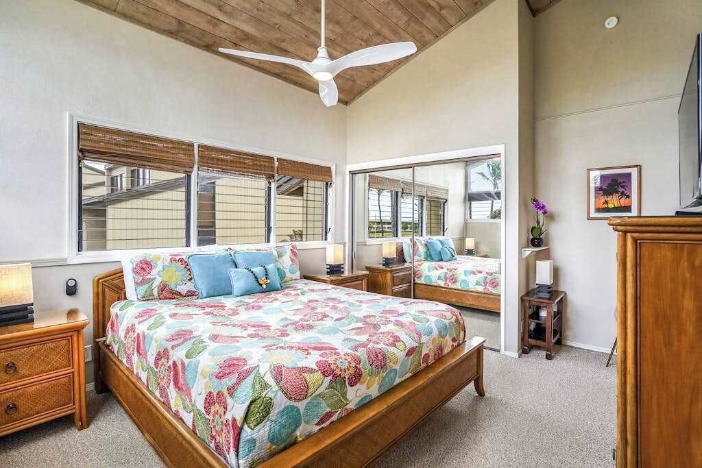 Un dormitorio con una cama grande, un ventilador de techo, una ventana con persianas, un espejo y una mesita de noche con lámpara.