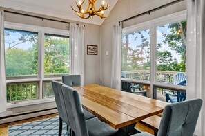 A dining area with a wooden table and grey chairs, large windows with a view of greenery, and a chandelier. - Beech on The Rocks | pet friendly, club access, mtn views (Banner Elk)