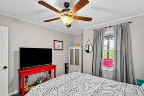 A bedroom with a bed, a television on a red table, a ceiling fan, a window with curtains, and a wall-mounted mirror. - Beech on The Rocks | pet friendly, club access, mtn views (Banner Elk)