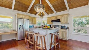 A kitchen with wooden flooring, a central island with stools, a refrigerator, and a hanging light fixture. - NEW Princeville Home for 12 with Mountain and Waterfall Views (Princeville)