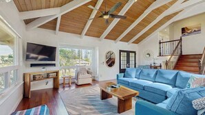 A spacious living room with a large TV, a blue sofa, wooden flooring, and a ceiling fan. - NEW Princeville Home for 12 with Mountain and Waterfall Views (Princeville)