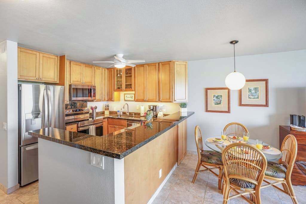 Una cocina con armarios de madera, una isla central, una zona de comedor con sillas de mimbre y un ventilador de techo.