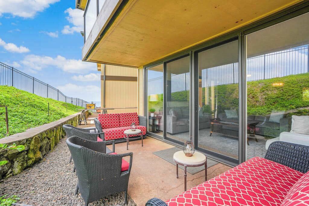 Een overdekt terras met rieten meubilair, een rode sofa met patroon en een glazen deur naar een patio.