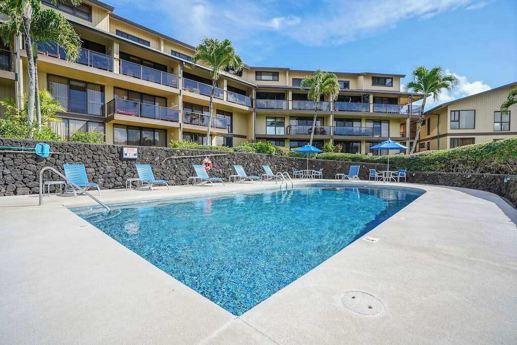 A swimming pool with lounge chairs, a stone wall, and a multi-story building with balconies.