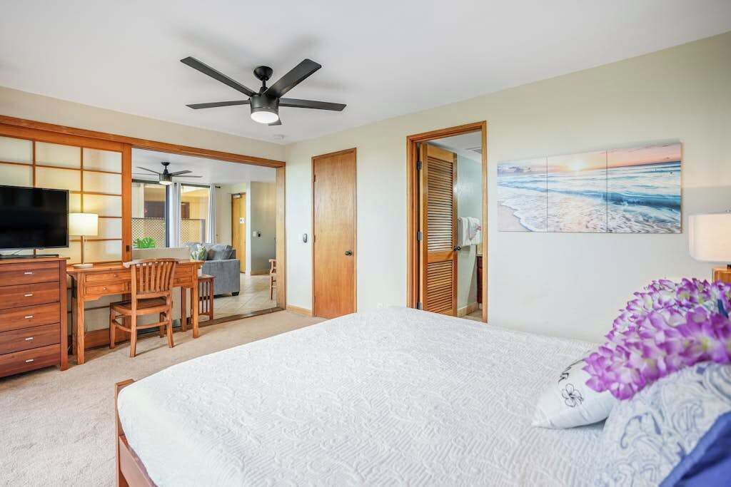 Habitación de hotel con cama, escritorio, silla, televisor, ventilador de techo y un cuadro con una escena de playa.
