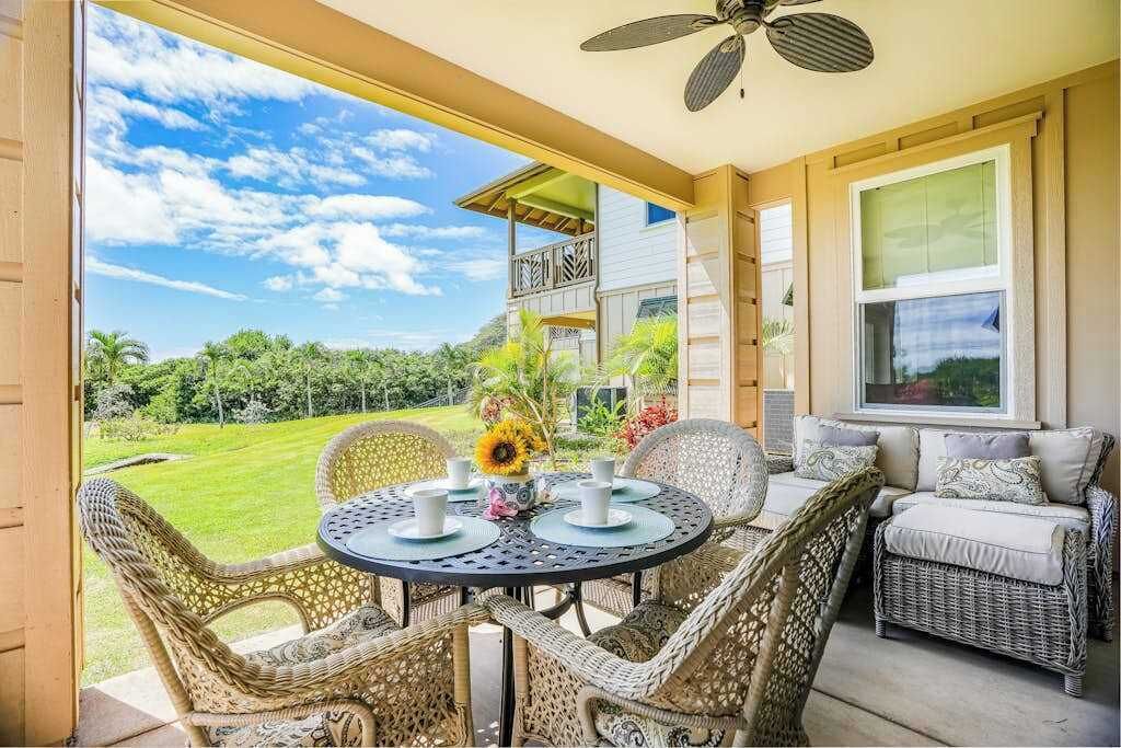 A covered patio with wicker furniture, a table with a vase of flowers, and a view of a house and landscape.