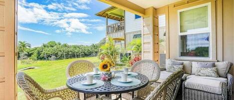 A covered patio with wicker furniture, a table with a vase of flowers, and a view of a house and landscape.