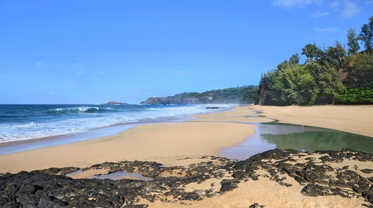 Ein Strand mit klarem Wasser, sandigem Ufer und einem felsigen Bereich im Vordergrund.