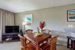 A dining area with a wooden table, chairs, a television, and artwork on the walls. - New Fall Special Kapaa 1BR with 3 Pools, 2 Hot Tubs and Beach Path (Kapaa)