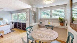 Ein Zimmer mit einem Bett, einem runden Holztisch mit Stühlen, einem Fenster mit Jalousien und einem kleinen Regal mit Gegenständen.