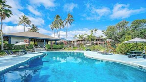 Une piscine avec des chaises longues, des palmiers et un espace abrité équipé de parasols.