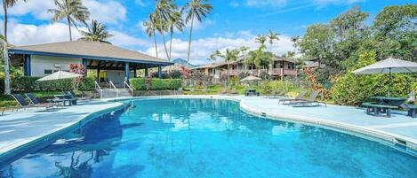 A swimming pool with lounge chairs, palm trees, and a covered area with umbrellas.
