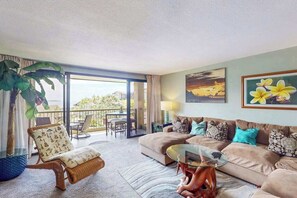 Ein Wohnzimmer mit einer Couch, einem Sessel und einem Glastisch. Ein Balkon bietet Blick ins Freie.
