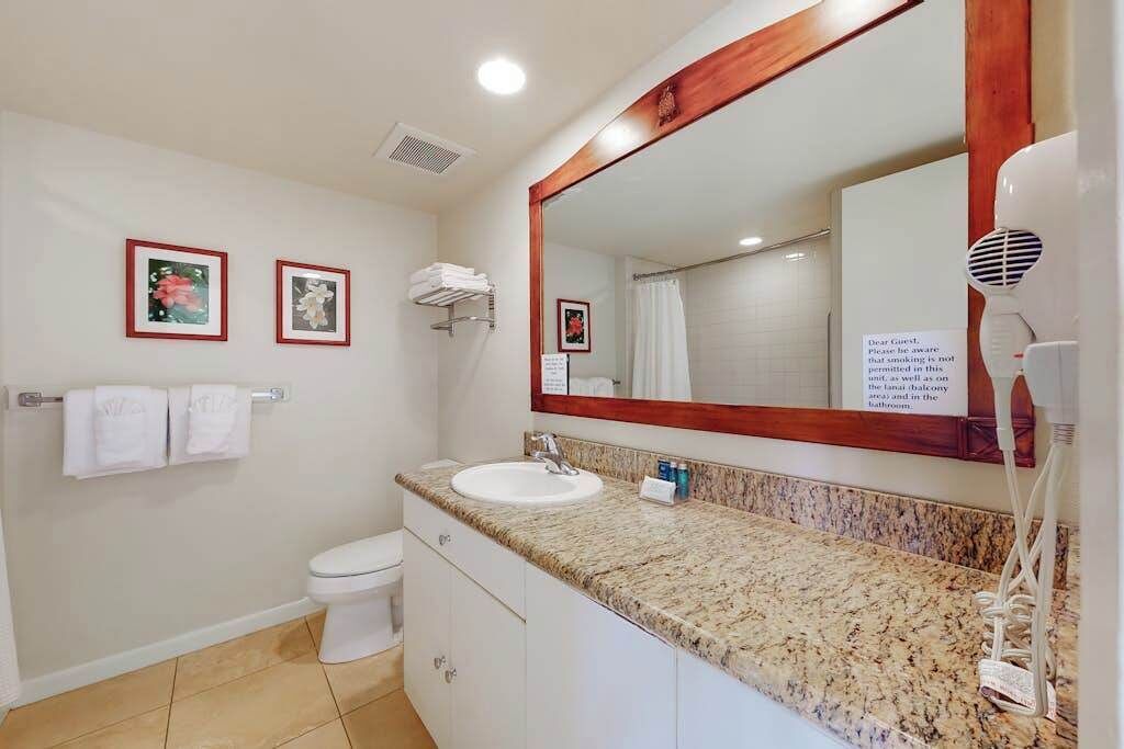 A bathroom with a sink, toilet, and a large mirror. There are framed pictures on the wall and a sign about no smoking.