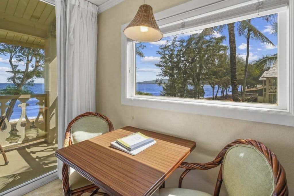 A room with a wooden table, two chairs, a hanging lamp, and a window with a view of palm trees and the sea.