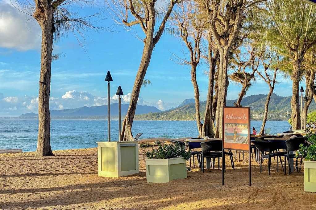 A beachside restaurant with outdoor seating, a welcome sign, and a view of the sea and mountains.