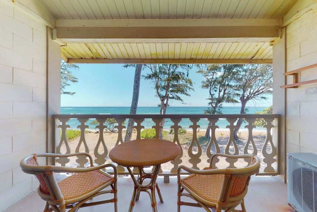 Un balcon abrité, doté de meubles en osier, offrant une vue sur la plage et s’ouvrant sur un ciel dégagé.