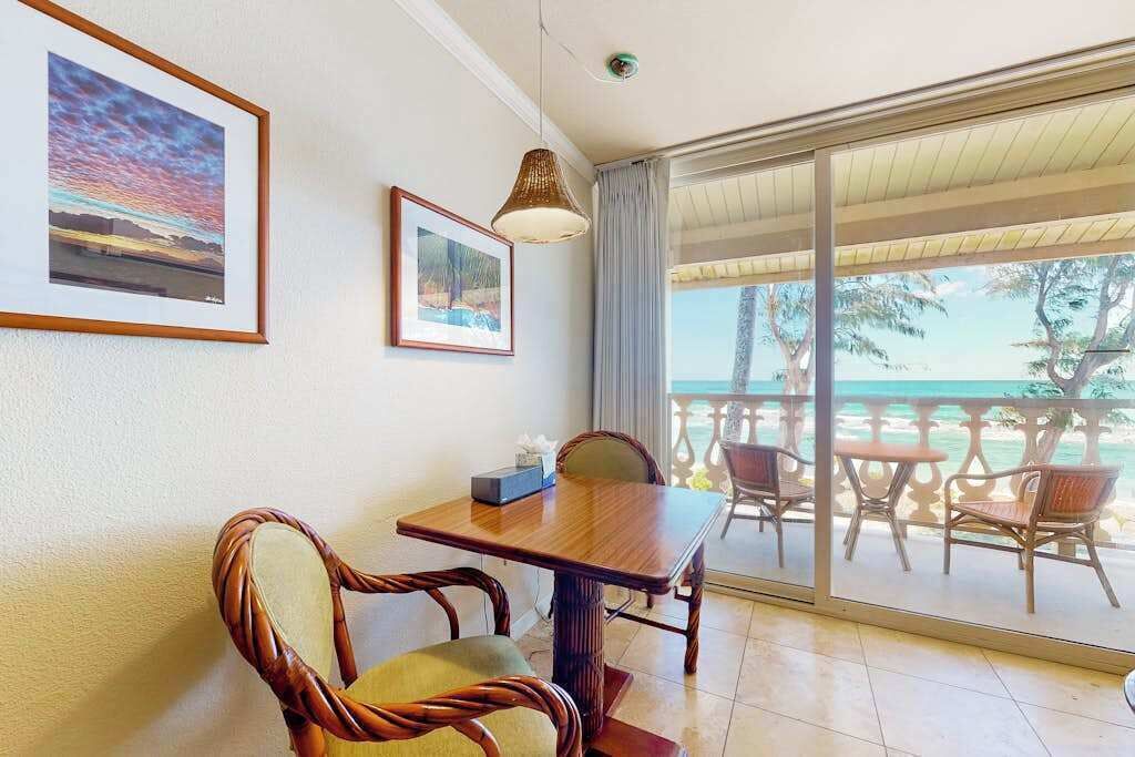 Una habitación con una mesa y sillas de madera, un ventanal con vista a la playa y cuadros enmarcados en la pared.