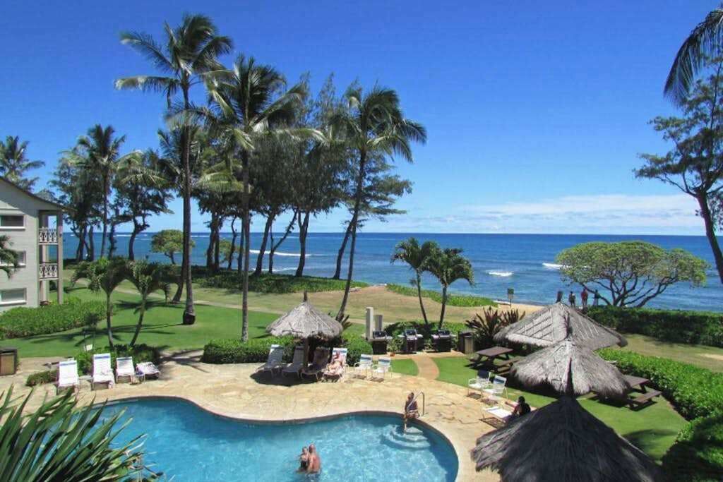 A resort pool with palm trees, a beach view, and a thatched-roof structure.