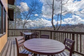 Eine Holzterrasse mit rundem Tisch und Stühlen, mit Blick auf eine gebirgige Landschaft.