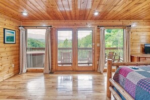 A wooden cabin with a large sliding door, a bed with a colorful quilt, and a view of a forested area. - Luxury Lookout (Gatlinburg)