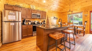 A wooden cabin interior with a kitchen featuring a refrigerator, microwave, and stove. There is a wooden island with stools and a Christmas tree. - Mimi and Pop's Place (Gatlinburg)