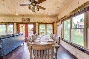 Uma sala de jantar com uma longa mesa de madeira posta para seis pessoas, rodeada por cadeiras de madeira, com vista para o exterior através de grandes janelas.