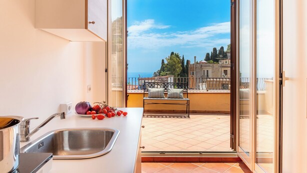 Eine Küche mit Spüle, Herd und Fenster mit Blick auf einen Balkon und Meer sowie Gebäude.