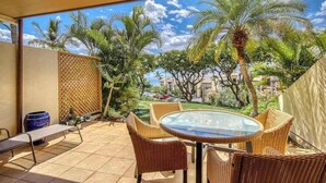 Un espacio exterior techado con mobiliario de mimbre, una mesa de vidrio y vistas a palmeras y edificios.