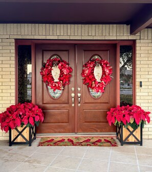 A double wooden door with two large wreaths, a doormat with bird designs, and two potted plants with red flowers. - Charming 3-bedroom house in Brownsville with cool AC breeze (Brownsville)