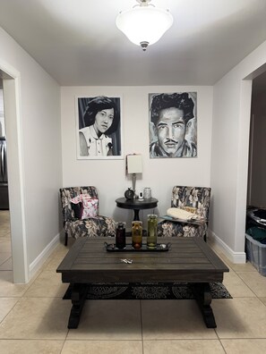 A room with a dark wooden coffee table, two floral-patterned armchairs, a small round table with a lamp, and two large framed portraits on the wall. - Charming 3-bedroom house in Brownsville with cool AC breeze (Brownsville)