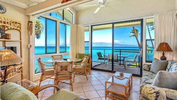 A spacious room with a large sliding glass door leading to a balcony with a view of the ocean. There are wicker furniture pieces, a table, and a sofa. - *NEW* Turtle-Watching Oceanfront Lanai in West Maui (Lahaina)