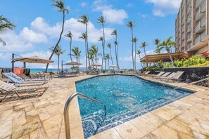 A swimming pool with lounge chairs and umbrellas, surrounded by palm trees and a hotel building. - Stunning Beachfront 2 Bedroom Condo in Kihei with Resort Amenities (Kihei)