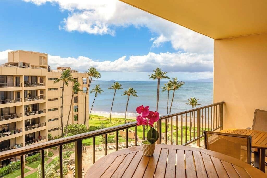 A balcony with a view of the ocean, palm trees, and a building in the background.