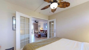 Ein Schlafzimmer mit einem Bett, einem Deckenventilator und Blick in einen Wohnbereich mit einer Couch und einer Lampe.