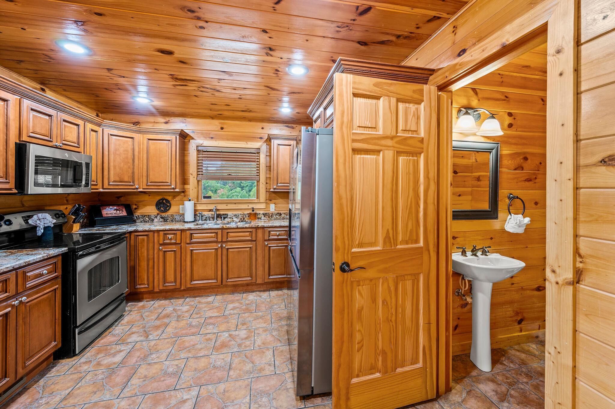 A wooden cabin kitchen with a stone floor, wooden cabinets, a microwave, stove, refrigerator, and a sink.