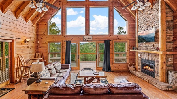 A log cabin interior with a fireplace, large windows, and a cozy seating area. - Mountain Wonderland (Sevierville)