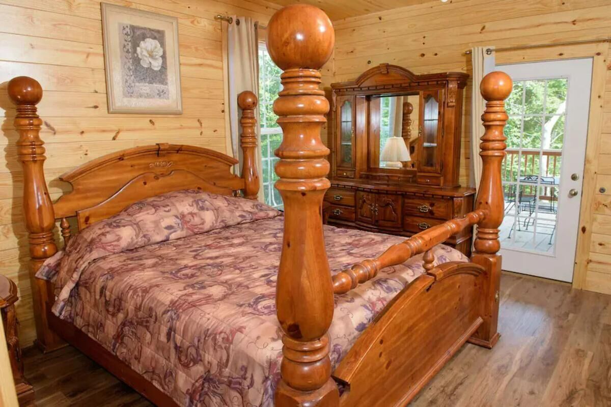 A wooden four-poster bed with a floral bedspread, a wooden dresser with a mirror, and a door leading to an outdoor area.