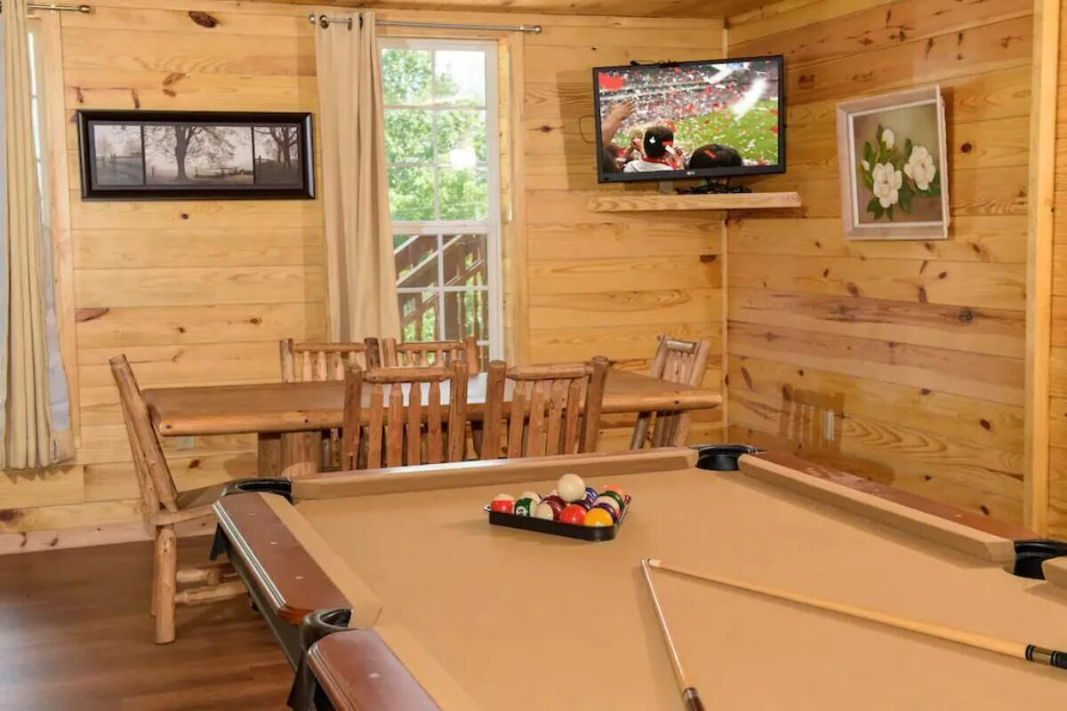 A wooden-paneled room with a pool table, a television mounted on the wall, and a dining table with chairs.