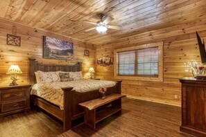 Quarto de cabana de madeira com cama, mesas de cabeceira, um banco, ventoinha no teto e janela com persianas.