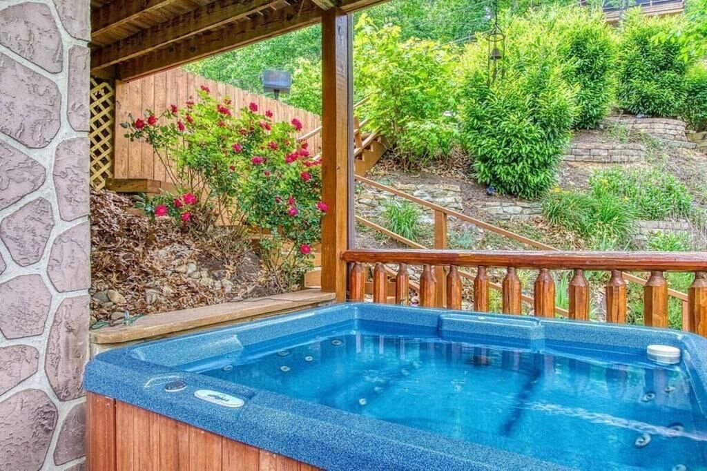 A hot tub with a stone wall and wooden deck, surrounded by greenery and a wooden fence.
