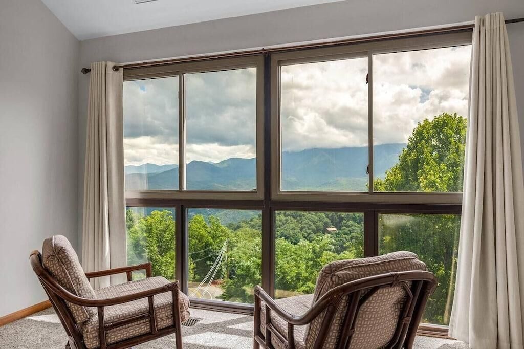A room with large windows offering a mountain view, two wicker chairs, and a carpeted floor.