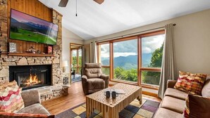 A living room with a stone fireplace, a television, a large window with a mountain view, and a ceiling fan. - Reed's Retreat (Gatlinburg)