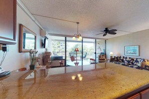 A spacious living area with a large kitchen island, a ceiling fan, and a view through large windows. - Kihei Condo with Pool and Beach Access (Kihei)